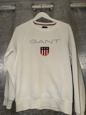 Vit sweatshirt från GANT med logga - Säljer en clean vit sweatshirt från GANT med broderad logga och sköld på bröstet. Tröjan har rund halsringning och långa ärmar. Perfekt till chill vardagsstil och funkar året runt.