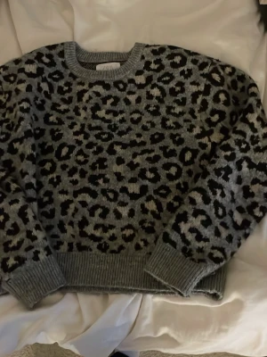 Grå leopardmönstrad stickad tröja - Säljer en stickad tröja från Zara med grå bas och leopardmönster i svart och beige. Tröjan har rund halsringning, ribbade muddar och är långärmad. Perfekt för dig som vill sticka ut lite extra i höst och vinter.