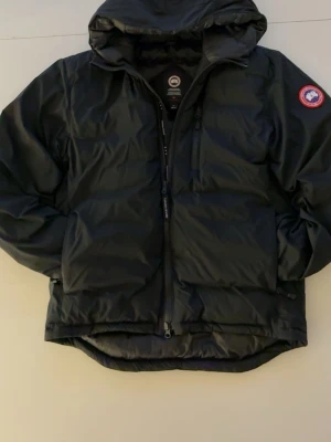 Svart dunjacka från Canada Goose - Säljer en svart dunjacka från Canada Goose med huva och klassisk logga på ärmen. Jackan har dragkedja framtill, två stora fickor och en clean look. Perfekt för kalla dagar och riktigt snygg till streetwear-stilen.