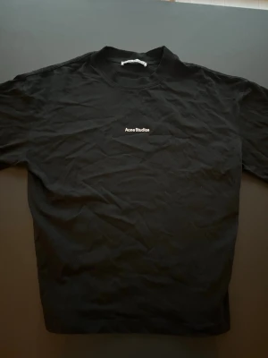 Acne Studios T-shirt Svart - Säljer en svart t-shirt från Acne Studios med enkel och clean design. T-shirten har korta ärmar och en liten vit logga broderad på bröstet. Tillverkad i mjuk bomull som känns skön mot huden. Perfekt till jeans eller shorts för en stilren look.