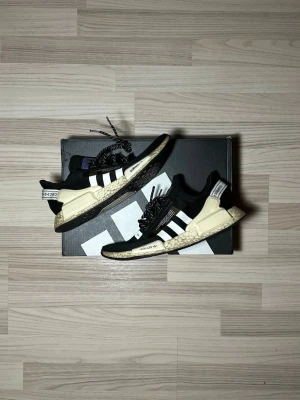 Adidas NMD_R1V2  sneakers svart/vit - Säljer ett par Adidas NMD_R1V2 sneakers i svart och vitt med klassiska tre ränder på sidan. Skorna har beige detaljer på mellansulan och hälen, samt svart/vita snören. Ovandelen är i textil och sula i syntet. Perfekta för dig som gillar streetwear och bekväma skor.