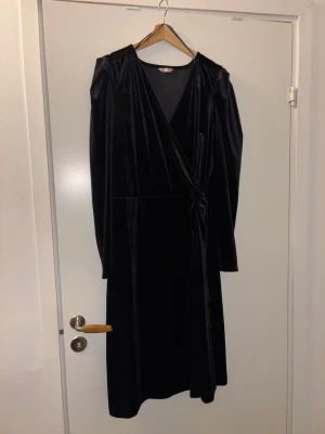 Svart omlottklänning i sammet 2XL - Svart långärmad omlottklänning i mjuk sammet med knyt i midjan. Klänningen har v-ringning och elegant fall. Perfekt för dig som vill ha en stilren och klassisk look med lite extra lyxig känsla.