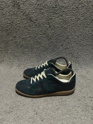 Masion margiela - Säljer ett par mörkblå sneakers med snygga mockadetaljer och beige snörning. Skorna har en klassisk låg profil, gummisula i brunt och inslag av både mocka och skinn. Perfekta för dig som gillar stilrena och tidlösa sneakers.