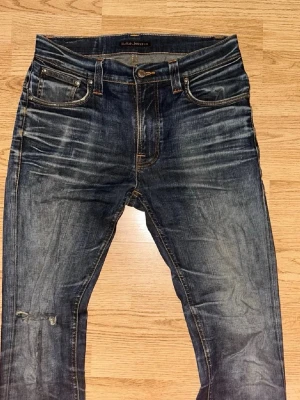 Nudie jeans blåa - Säljer ett par nudie  jeans i storlek w31 L34 mörkblå tvätt med snygga slitningar och whiskers. Klassisk femficksmodell med orange sömmar och låg midja. Jeansen har en smal passform och är tillverkade i slitstarkt denimtyg. Perfekta för dig som gillar en avslappnad men trendig look.