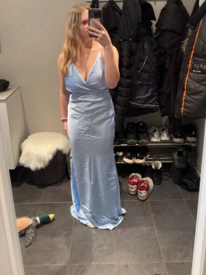 Ljusblå långklänning med öppen rygg - Säljer en elegant ljusblå långklänning med djup v-ringning och smala axelband som korsas i ryggen. Klänningen har öppen rygg och knyt bak, vilket ger en snygg och modern look. Perfekt för speciella tillfällen och sommarens fester. Prislapp finns kvar. Säljer då den var för stor.