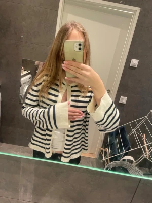 Randig kofta med guldknapp - Säljer en vit och svart randig kofta med lång ärm och snygg guldknapp vid halsen. Koftan är stickad och har en loose passform, perfekt att slänga på sig över en t-shirt. Klassisk och stilren look som funkar året runt.