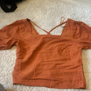 Rostorange blus med puffärm - Säljer en snygg rostorange blus med fyrkantig ringning och puffiga korta ärmar. Blusen har ett korsband i ryggen och en smockad detalj baktill som ger en cool form. Materialet känns lätt och mjukt, perfekt för vår och sommar. Passar dig som gillar färg och trendiga detaljer.