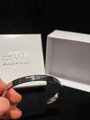 Maison Margiela silverfärgad armband - Säljer ett stilrent och minimalistiskt armband från Maison Margiela i silverfärg. Armbandet är öppet och har Maison Margiela Paris ingraverat framtill. Kommer med original ask och är perfekt för dig som gillar clean design.