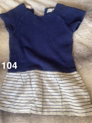 Marinblå stickad tunika med ränder - Säljer en söt marinblå stickad tunika med korta ärmar och rund hals. Nedre delen är vit med tunna blå ränder för en fräsch look. Perfekt till jeans eller kjol för en avslappnad stil🌸 .En liten fläck. Storlek 104