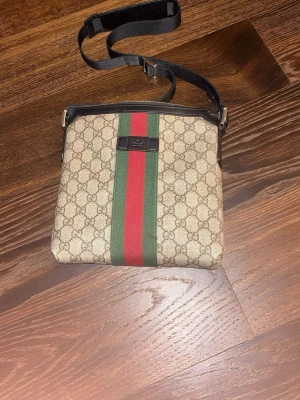 Gucci axelväska med GG-mönster - Säljer en snygg axelväska från Gucci med klassiskt GG-mönster i beige och brunt. Väskan har en bred grön och röd rand i mitten och justerbar svart axelrem. Perfekt för dig som vill ha en ikonisk och stilren accessoar.
