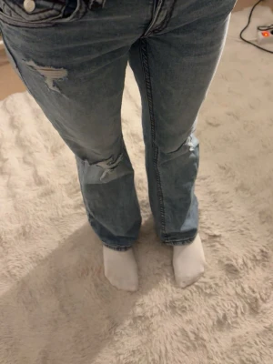 Blå bootcut jeans med slitningar - Säljer ett par ljusblå bootcut jeans med slitna detaljer vid knäna och låret. Jeansen har låg midja och klassiska fickor med knappdetalj bak. Materialet är jeans i bomull och passformen är loose med en avslappnad vibe.