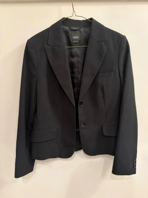 Svart kavaj från Esprit - Snygg svart kavaj från Esprit med klassisk krage, två knappar framtill och fickdetaljer. Figurnära passform och helfodrad insida. Perfekt till både jeans och kostymbyxor för en clean look.