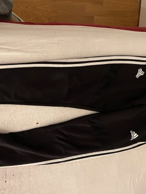 Svarta Adidas träningsbyxor med vita ränder - Säljer ett par svarta träningsbyxor från Adidas med klassiska vita ränder längs sidorna och Adidas-logga på benet. Byxorna har elastisk midja och är gjorda i ett mjukt syntetmaterial som passar perfekt till träning eller chill. Snygg och sportig look!