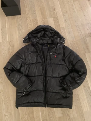 Svart pufferjacka från Polo Ralph Lauren - Säljer en svart pufferjacka från Polo Ralph Lauren med huva och dragkedja. Jackan har en röd logga på bröstet och två fickor med dragkedja. Perfekt för kalla dagar och riktigt snygg till streetwear-stilen. Väldigt bra skick bara använd i december!