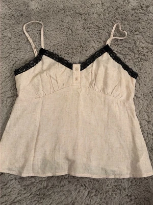 Beige linnetopp med spets från SHEIN - Säljer en beige linnetopp från SHEIN med tunna axelband och svart spetsdetalj längs urringningen. Toppen har två små knappar framtill och är tillverkad i ett luftigt material som känns perfekt för varma dagar.