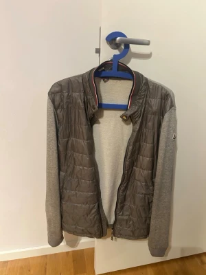 Äkta Grå Moncler cardigan - Säljer en grå Moncler jacka med quiltad front i mörkgrått och ärmar i mjuk bomull. Jackan har ribbad krage med röd och vit detalj, dragkedja framtill och Moncler-logga på ärmen. Skicka gärna prisförslag! Finns några små defekter men absolut ingen som påverkar. Är storlek L men passar mindre, är 180 och den passar bra. Självklart äkta och finns äkthetsbevis!