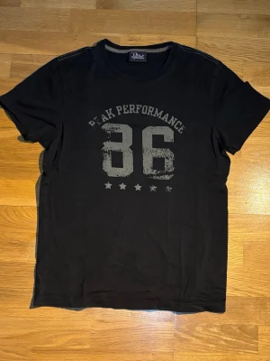 Svart Peak Performance t-shirt - Svart t-shirt från Peak Performance med tryck '86' och texten 'Peak Performance' i grått framtill. Klassisk rund halsringning och korta ärmar. Tillverkad i mjuk bomull för skön känsla. Perfekt för en avslappnad och sportig stil.