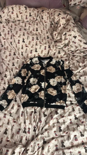 Blommig bomberjacka från Happy Holly - Säljer en svart bomberjacka från Happy Holly med stora beige och vita blommor. Jackan har ribbade muddar vid ärmslut och hals, samt dragkedja framtill. Perfekt för dig som vill sticka ut med ett snyggt blommönster och sportig känsla.