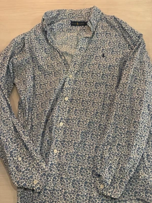 Blommig skjorta från Ralph Lauren - Säljer en snygg skjorta från Ralph Lauren med blått blommönster på vit botten. Skjortan har klassisk krage, långa ärmar och knäppning framtill. Tillverkad i mjuk bomull som känns skön mot huden. Perfekt för dig som vill sticka ut lite extra.