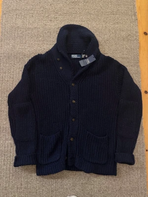 Mörkblå stickad kofta Polo Ralph Lauren - Säljer en mörkblå stickad kofta från Polo Ralph Lauren. Lägg märke till att tröjan är medvetet distressed för att ge en cool vintage look. Nypris cirka 8000 kr. Koftan har grovstickad struktur, stora fickor framtill, knappar hela vägen och en rejäl krage. Materialet är 55% linne och 45% bomull. Perfekt för lager-på-lager under kyliga dagar. Det är storlek amerikansk S vilket motsvarar M i Europa.