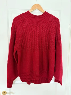 Röd kabelstickad tröja med rund hals - Mysig röd kabelstickad tröja med bred ribbad rund halsringning och långa ärmar. Tröjan har ett snyggt kabelmönster upptill och ribbade muddar vid ärmslut och nederkant. Perfekt för kyliga dagar och enkel att styla till jeans eller kjol.