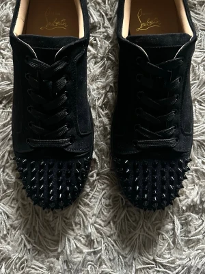 Christian Louboutin  - Säljer ett par svarta sneakers från Christian Louboutin i mocka med ikoniska röda sulor. Skorna har svarta nitar över hela tån, svarta skosnören och en stilren, låg siluett. Perfekta för dig som vill sticka ut med exklusiva detaljer. Skorna är storlek 43.