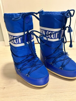 Blå Moon Boots med snörning - Säljer ett par supersnygga  blå Moon Boots med vit logga och snörning runt hela skaftet.  Aldrig använda !  Storlek 35-38 