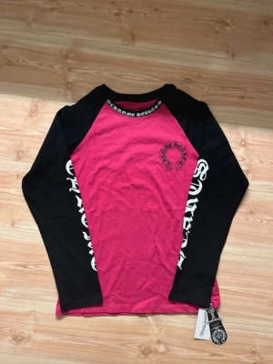Chrome hearts longsleeve  - Säljer en långärmad topp från Chrome Hearts med svart tryck på bröstet och ryggen. Toppen har svart text på ärmarna och kragen, samt kontrast mellan rosa kropp och svarta ärmar. Tillverkad i mjuk bomull och har en avslappnad passform.