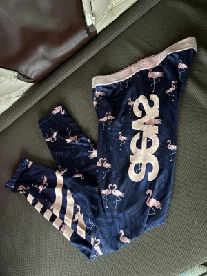 Mönstrade leggings från Svea - Säljer ett par snygga leggings från Svea i mörkblått med rosa flamingomönster och stora Svea-tryck på benet. Midjebandet är ljusrosa och byxorna har coola ränder på ena benet. Perfekta för dig som vill sticka ut med färg och mönster.