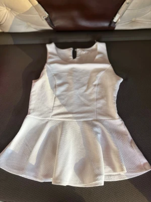 Vit peplumtop från Jennifer Taylor - Säljer en vit peplumtop från Jennifer Taylor i storlek M. Toppen är ärmlös med rund halsringning och har en strukturerad, lite tjockare textur. Den har en snygg utsvängd nederdel som ger en peplumeffekt och en liten knappdetalj i nacken.