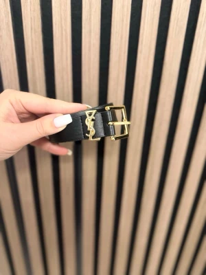 Ingen väst på bilden - Bilden visar ett svart bälte med gulddetaljer och YSL-logga, ingen väst syns.