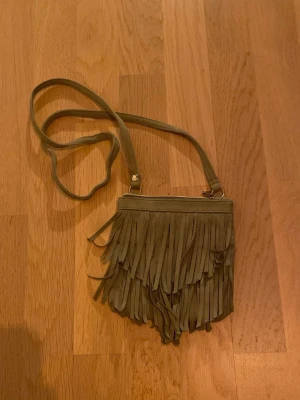 Beige axelväska med fransar - Säljer en grön  axelväska i mocka-look med långa fransar på framsidan. Väskan har smal axelrem och stängs med dragkedja upptill. 
