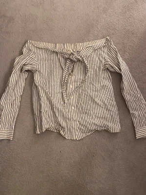 Randig offshoulder blus med knyt - Säljer en vit och mörkblå randig offshoulder blus med långa ärmar och knyt framtill. Blusen har knappar längs framsidan.