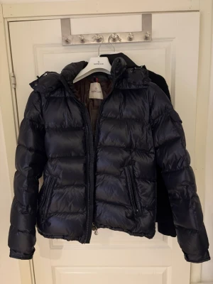 Moncler maya navy - Säljer en svart pufferjacka från Moncler med glansig finish och klassisk quiltad design. Jackan har huva, dragkedja framtill och två snedställda fickor med dragkedja. Insidan har en unik Moncler-serieetikett och innerficka med dragkedja. Perfekt för kalla dagar. Storlek 3