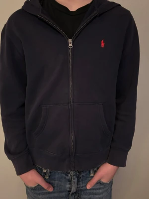 Mörkblå polo Ralphlauren kofta 14-16 år -  Mörkblå huvtröja från Polo Ralph Lauren i klassisk modell med hel dragkedja framtill. Tröjan har huva utan snörning, två öppna framfickor samt det ikoniska broderade Polo-emblemet i rött på bröstet. Insidan är mjuk och borstad för extra komfort. 	•	Märke: Polo Ralph Lauren 	•	Färg: Mörkblå 	•	Modell: Huvtröja med dragkedja 	•	Storlek: L (14–16), motsvarar barn/ungdom 	•	Tillverkad i: Kambodja 	•	Passform: Klassisk 	•	Material: Bomullsblandning (typisk sweatkvalitet)  En bekväm och tidlös hoodie