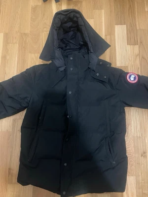 Svart dunjacka från Canada Goose - Säljer en svart Canada Goose dunjacka med huva och klassisk logga på ärmen. Jackan har dragkedja och knappar framtill samt två stora fickor. Medföljer avtagbar pälsdetalj till huvan och originalpåse. Perfekt för kalla vinterdagar.