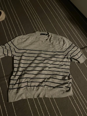 Grå randig tröja med kort ärm - Säljer en grå tröja med svarta horisontella ränder. Tröjan har korta ärmar och rund halsringning. Tillverkad i ett tunt och mjukt material, perfekt för vår och sommar. Enkel och stilren design som passar till det mesta. Storlek L men känns mindre