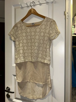 Beige spetsblus från Vero Moda - Supersöt beige blus från Vero Moda med korta ärmar och snyggt spetsmönster upptill. Toppen har en längre, tunnare underdel i samma färg som ger en cool lager-på-lager-look. Perfekt till jeans eller kjol för en trendig stil.