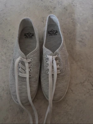 Ljusgrå sneakers med snörning, strl 39 - Säljer ett par ljusgrå sneakers i storlek 39. Skorna har klassisk rund tå, vita snören och platt sula. Tillverkade i mjukt tygmaterial som ger en clean och enkel look. Perfekta för dig som gillar minimalistisk stil.