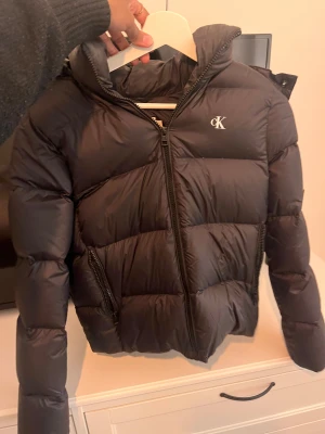 Svart pufferjacka från Calvin Klein - Säljer en svart pufferjacka från Calvin Klein med huva och dragkedja framtill. Säljer den då den inte är till användning. Den är inte använd ofta och ser nästan helt ny ut 
