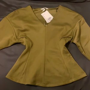 Olivgrön v-ringad topp från H&M - Snygg olivgrön topp från H&M med v-ringning och markerad midja, peplummodell. Toppen är helt ny med prislappen på, säljer då den inte kom till användning!