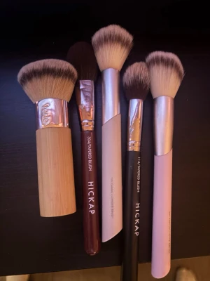 Fem sminkborstar från Hickap och andra - Säljer ett set med fem sminkborstar i olika storlekar och former, perfekta för applicering av puder, rouge och bronzer. Borstarna har mjuka syntetiska strån och handtag i trä, vinrött, svart och pastellrosa. Två av borstarna är märkta med Hickap.