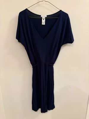 Mörkblå kortklänning från Alma Ras - En stilren mörkblå kortklänning från Alma Ras med v-ringning och knytband i midjan. Klänningen har korta ärmar och är tillverkad i ett mjukt, lätt material som känns skönt mot huden. Perfekt för dig som gillar enkel och snygg design.