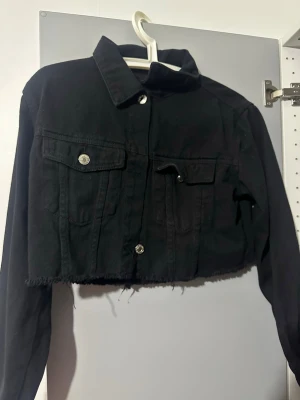 Svart croppad jeansjacka - Säljer en svart croppad jeansjacka med rå kant nertill och silvriga knappar. Jackan har två bröstfickor med lock och klassisk krage. Perfekt för dig som vill ha en edgy och trendig look.