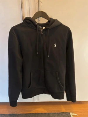 Svart Zip Up från Polo Ralph Lauren - Klassisk svart hoodie från Polo Ralph Lauren med dragkedja framtill och vit broderad logga på bröstet. Tröjan har huva med snörning, två fickor och ribbade muddar. Perfekt för en chill och stilren look.