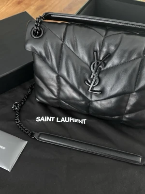 Svart quiltad axelväska från Saint Laurent - Säljer en svart quiltad axelväska från Saint Laurent med ikonisk YSL-logga framtill. Väskan är tillverkad i mjukt skinn och har en kedjerem med läderdetalj. Perfekt för dig som vill ha en stilren och lyxig accessoar.