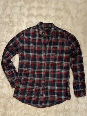 Rutig skjorta från Berga i bomull - Säljer en klassisk rutig skjorta från Berga i mörkblått, rött och vitt. Skjortan har långa ärmar, knappar framtill och en bröstficka. Tillverkad i 100% bomull för en skön känsla och avslappnad look.