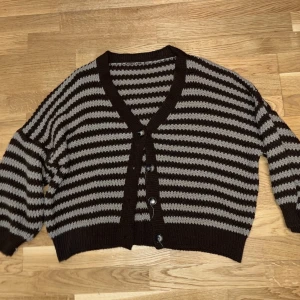 Randig stickad kofta i brunt och grått - Säljer en mysig stickad kofta med bruna och gråa ränder. Koftan har v-ringning, knappar framtill och ribbade muddar vid ärmslut och nederkant. Perfekt för lager på lager och chill dagar.