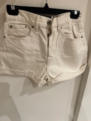 Vita high waist shorts från STYLE:DIBA - Säljer ett par vita high waist shorts från STYLE:DIBA i klassisk femficksmodell. De har uppvikta benslut, bälteshällor och stängs med knappar framtill. Materialet är kraftig bomull som ger en snygg och avslappnad look.