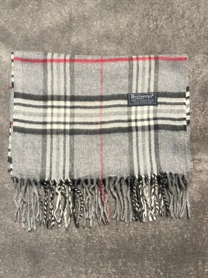 Burberry halsduk i grå - Klassisk grå halsduk från Burberrys i 100% lammull. Rutigt mönster i grått, svart, vitt och röda detaljer. Tidlös design med tvinnade fransar i ändarna och en snygg Burberrys-label. Perfekt accessoar för kyliga dagar.